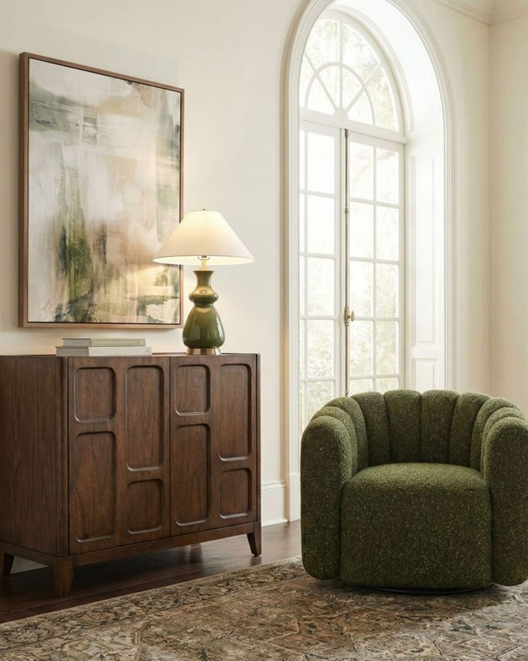 Cozy corner with a green upholstered chair, wooden cabinet, table lamp, and large arched window. 
