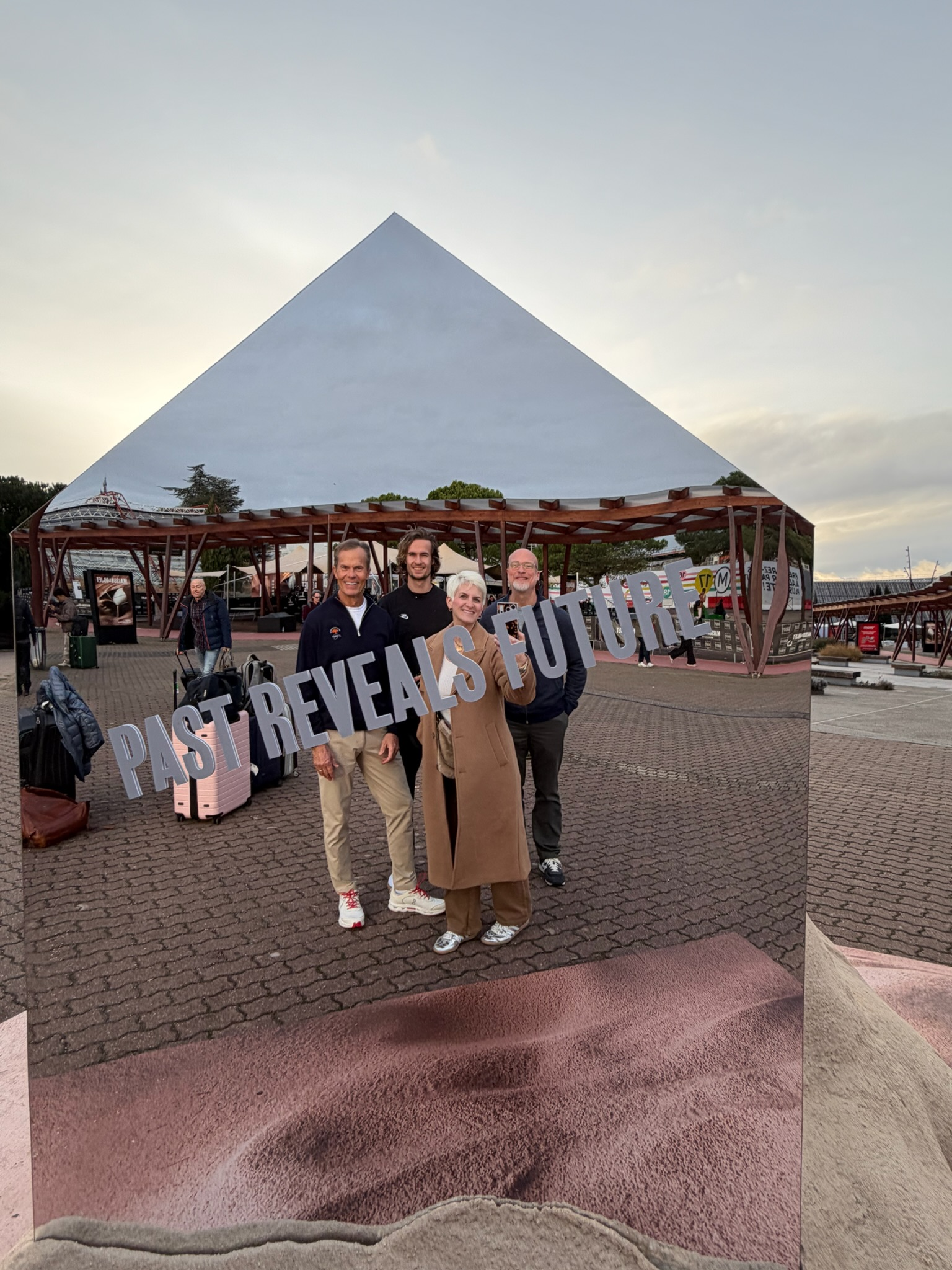 Four people posing in front of a mirrored intallation and pyramid-shaped building with "Past Reveals Future" text