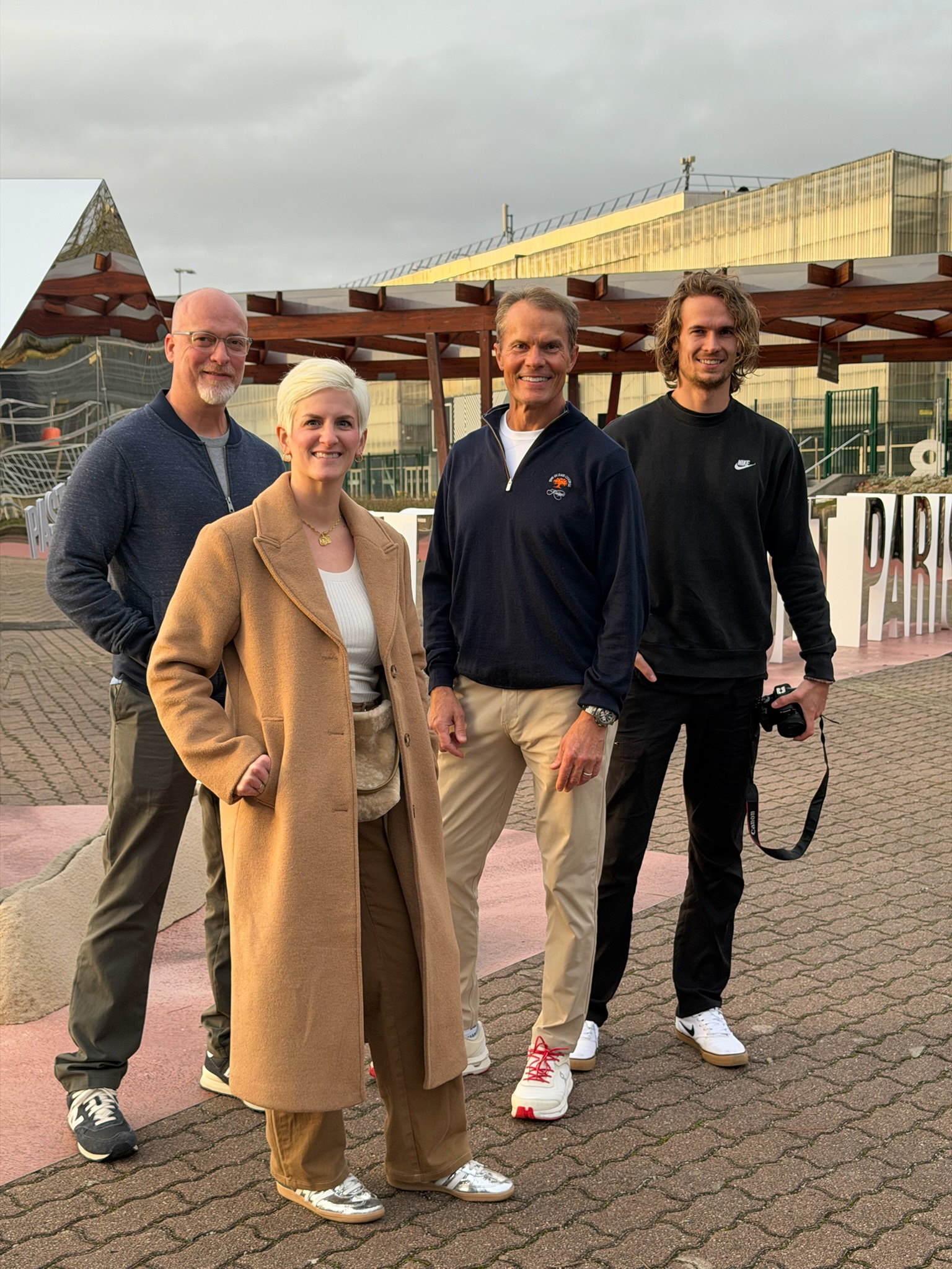 Four people posing together outdoors near a mirrored pyramid installation and modern buildings