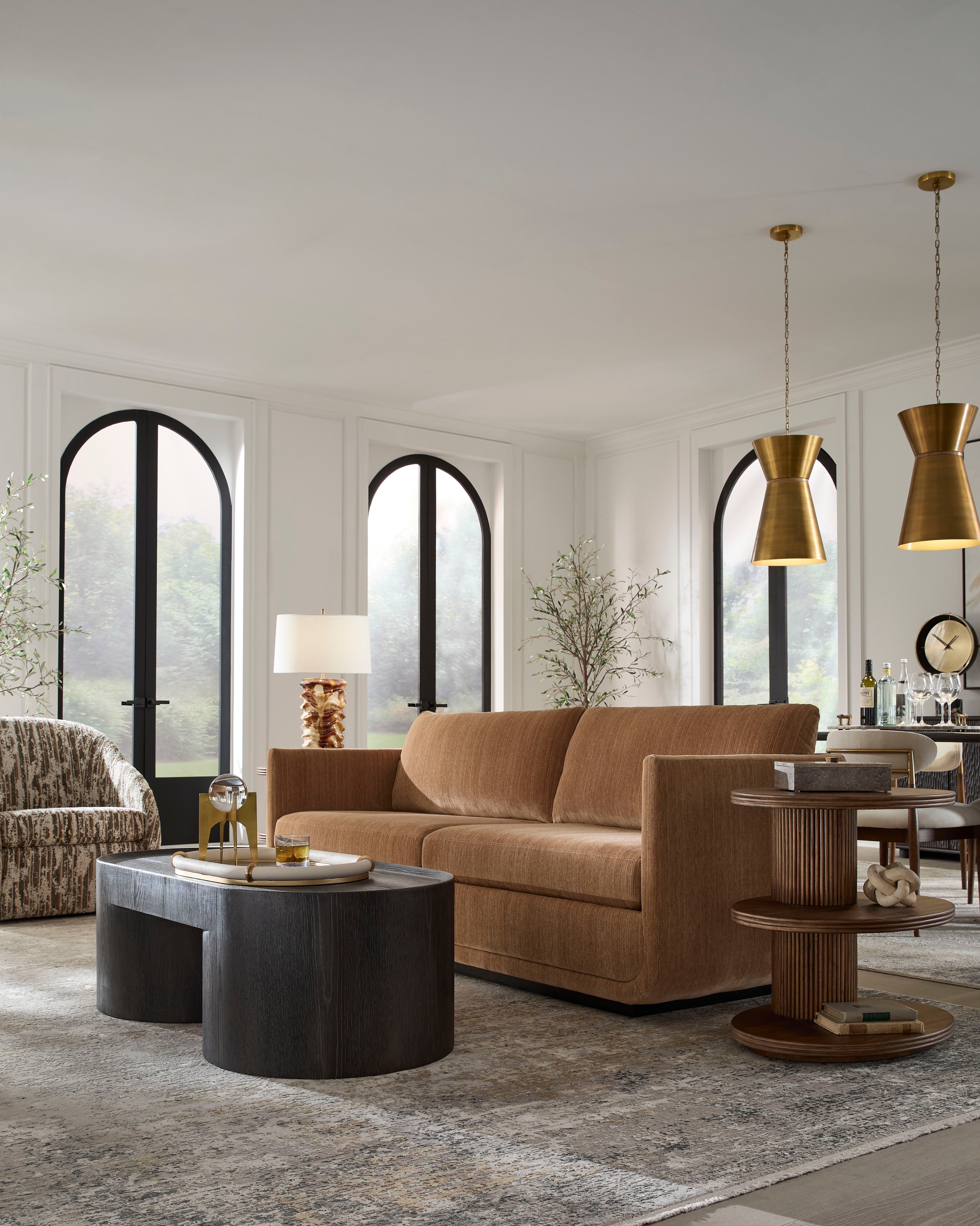Modern living room with a tan sofa, round wood tables, brass pendant lights, and tall arched windows.
