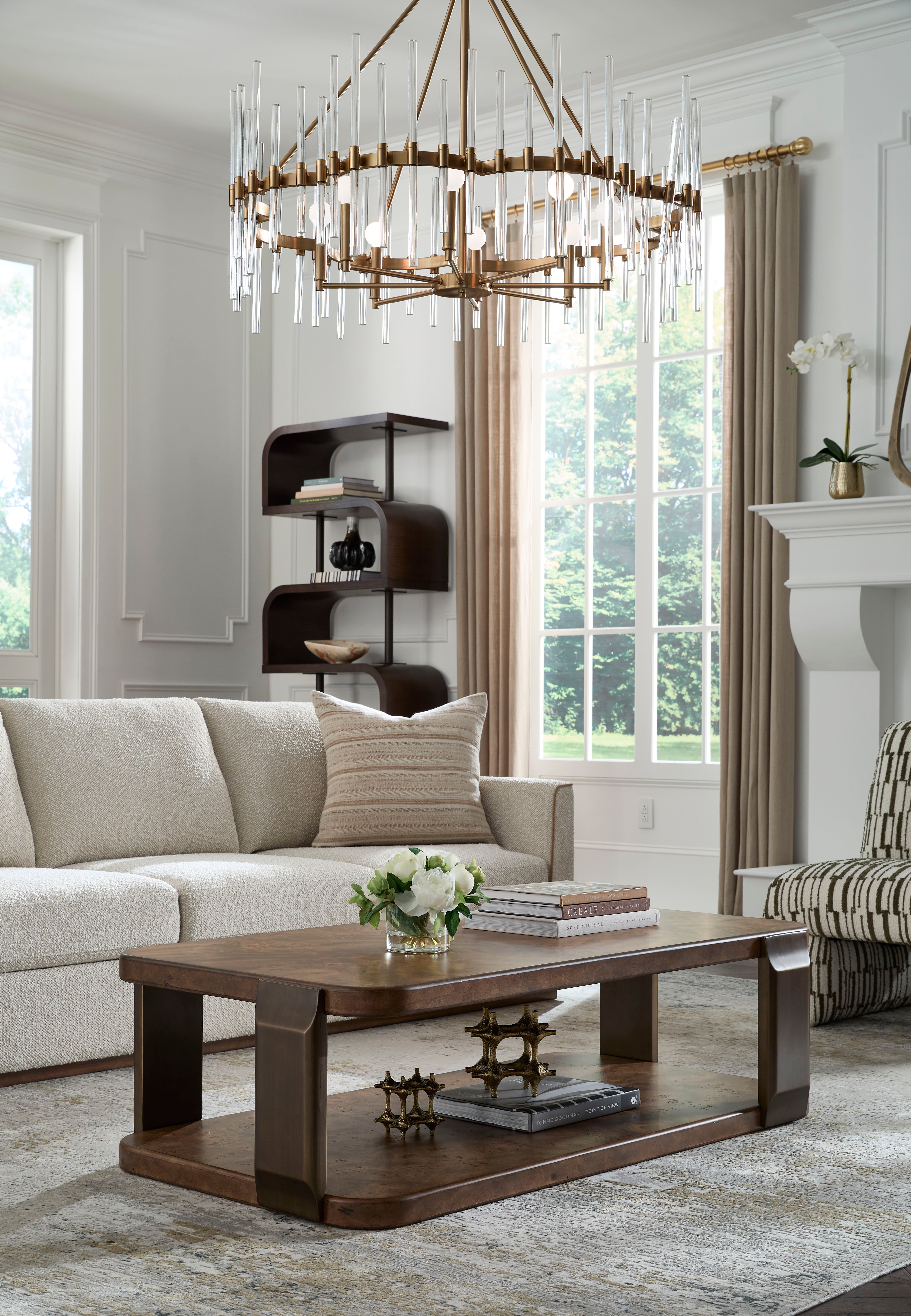 A living room featuring a large brass chandelier above a wooden coffee table. The space includes a light-colored sofa with a neutral accent pillow and a patterned armchair.