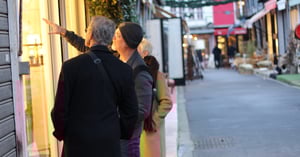 Two people standing on a quiet street, looking at and pointing toward a warmly lit shop window display 