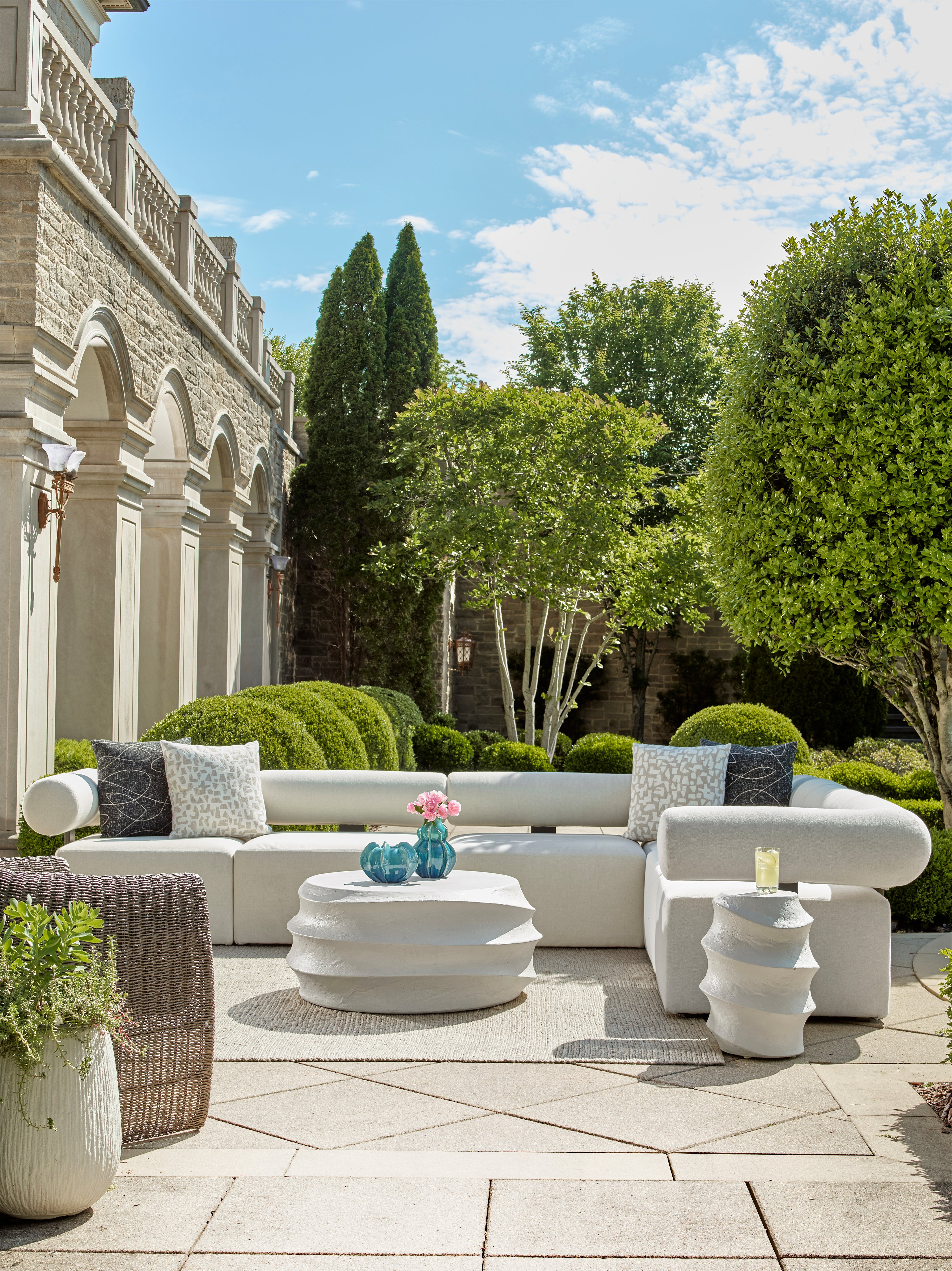 Outdoor patio with a white sectional sofa and sculptural coffee tables set against a landscaped garden