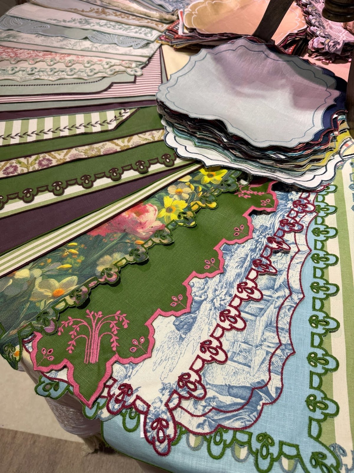 Stacked decorative placemats and linens with colorful patterns and scalloped embroidered edges