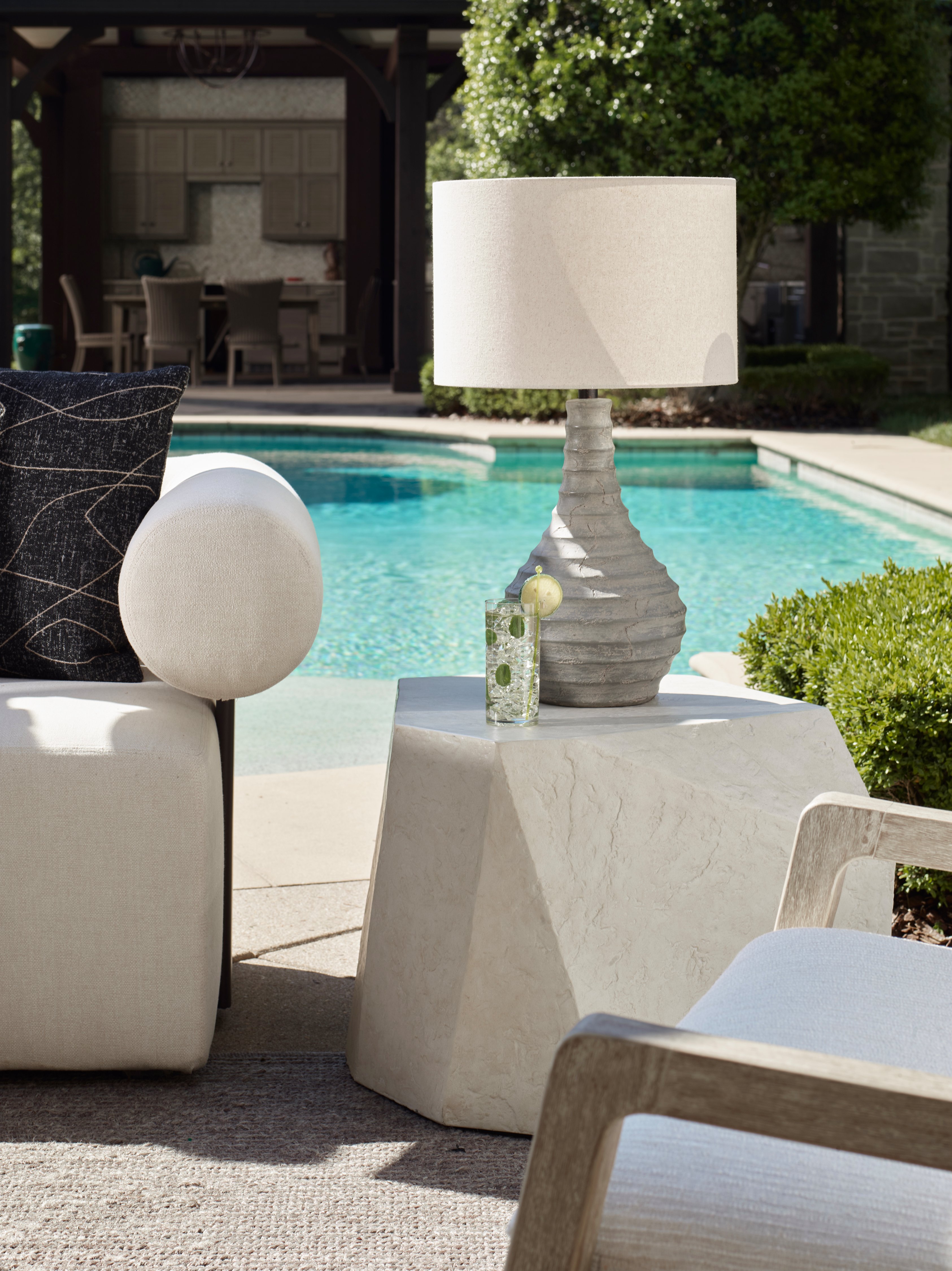 Outdoor side table with a textured gray lamp and drink beside a poolside seating area